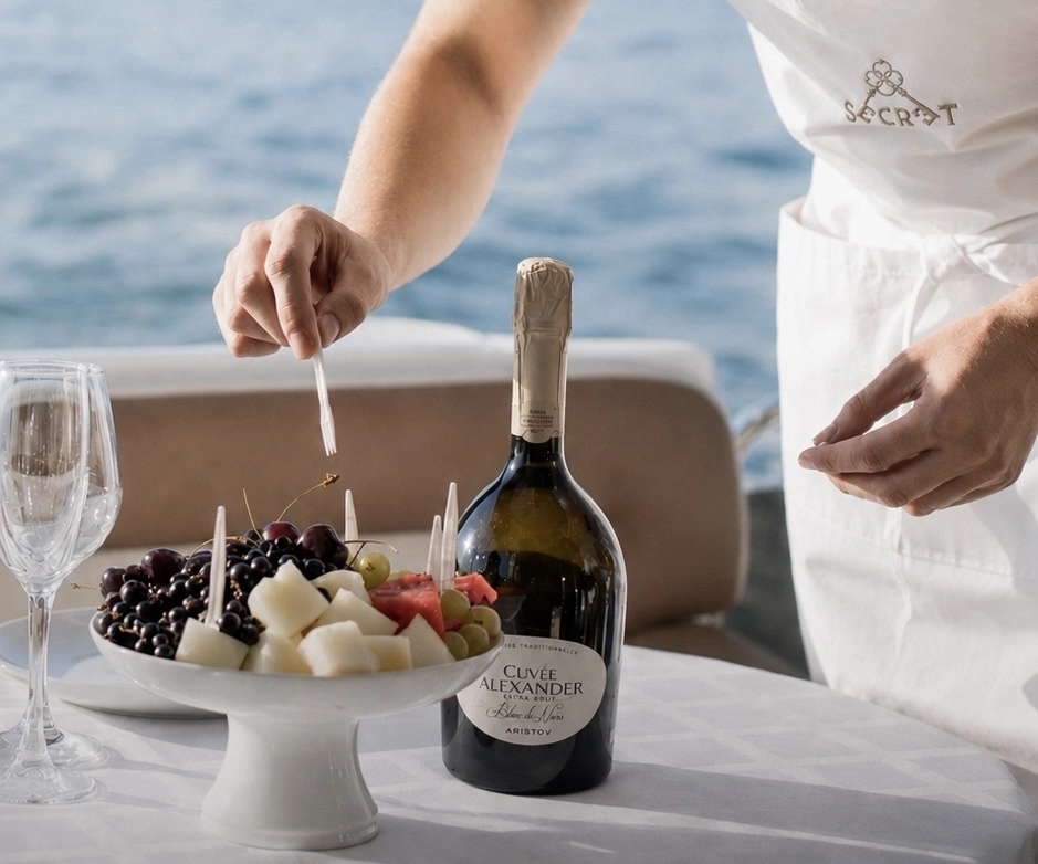 Crew member in a Secret Waves apron plating a fruit platter beside chilled sparkling wine on board in Dubai
